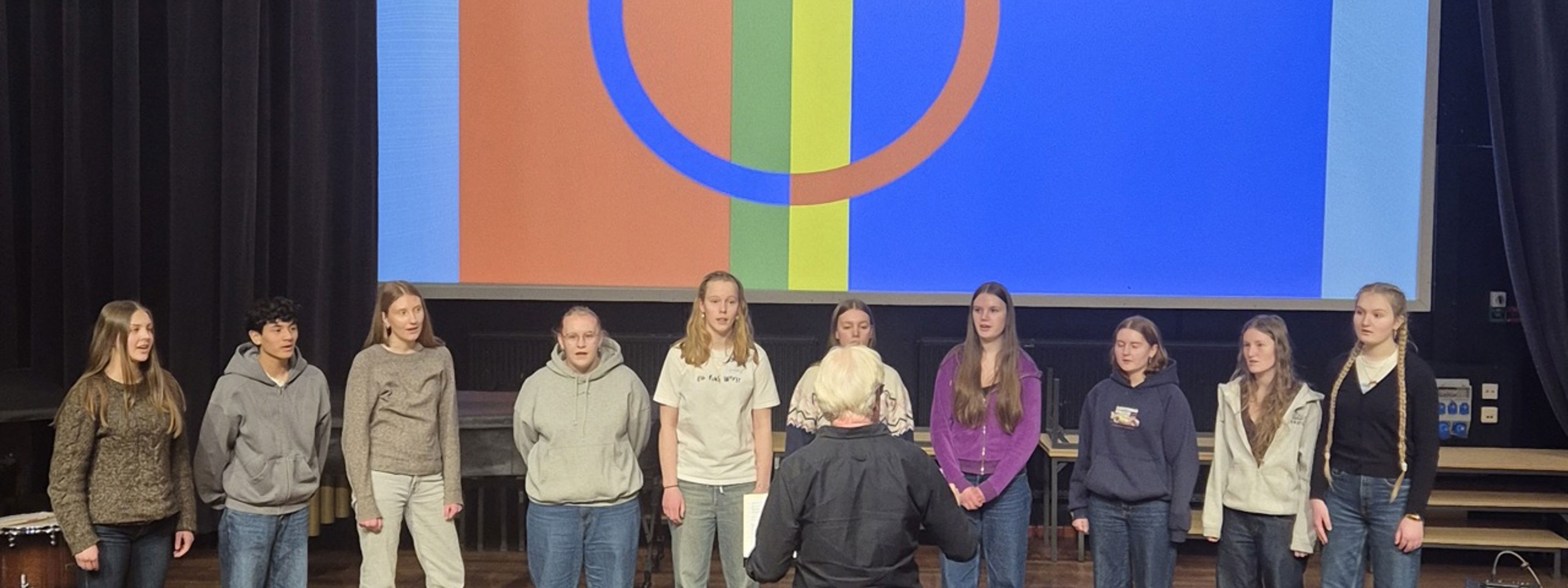 Elevar på musikklinja som syng samiske songar framfor eit bilete av samane sitt flagg